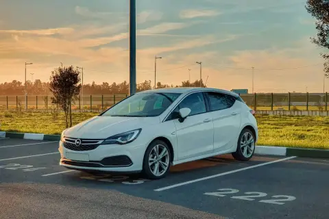 Biała Opel Astra K zaparkowana na parkingu. Słońce zachodzi, tworząc ciepłą atmosferę. Czas na opel astra k wymiana akumulatora.