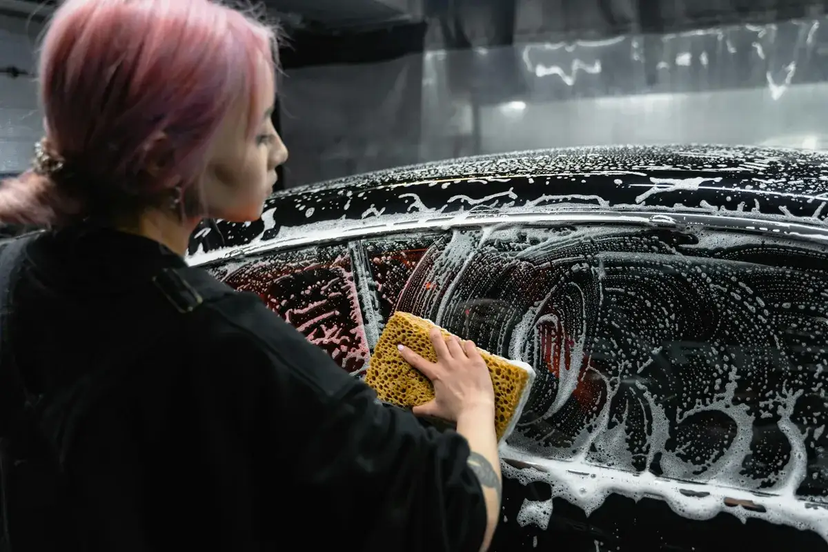 Kobieta z różowymi włosami myje auto gąbką. Pytanie: czym umyć szyby w aucie żeby nie parowały?